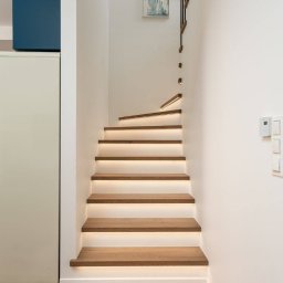 PRE-LOVED - Nowoczesne schody drewniane z podświetleniem LED, białe ściany, drewniana podłoga, minimalistyczny design, zegar na ścianie, obraz w tle.
