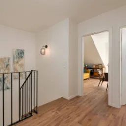 Nowoczesne wykończenie wnętrza: drewniana podłoga, czarna balustrada, abstrakcyjne obrazy i szklane kinkiety na białych ścianach, z widokiem na pokój na poddaszu.