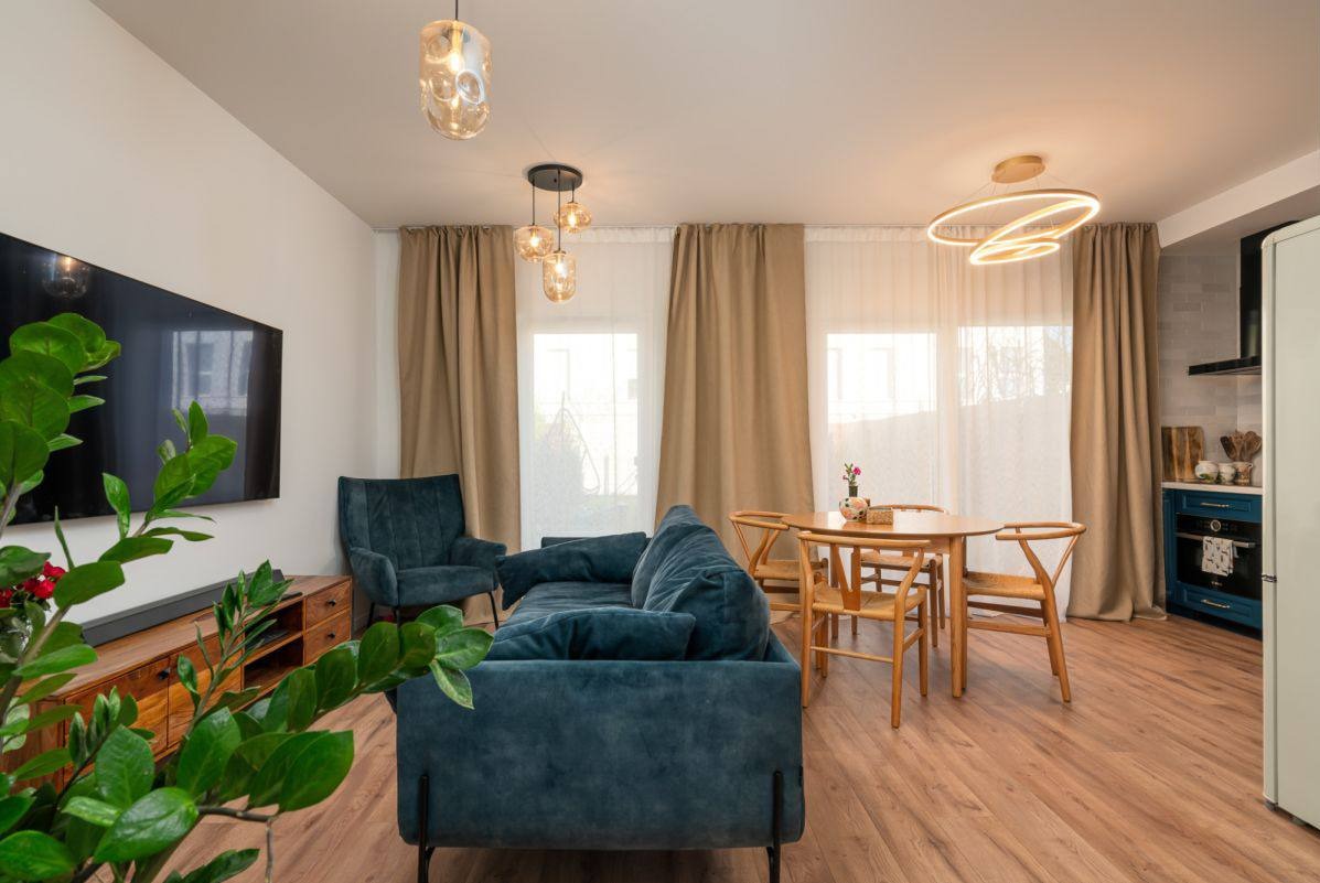 Stylowe wnętrze: turkusowa sofa, drewniany stół z krzesłami, designerskie lampy wiszące, drewniana podłoga i szafka RTV z rośliną. Nowoczesny salon z elementami vintage.
