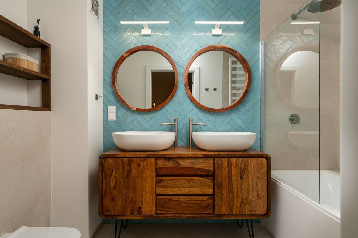 Nowoczesna łazienka z drewnianą szafką pod umywalki, okrągłymi lustrami i błękitnymi płytkami ułożonymi w jodełkę. Widoczna kabina prysznicowa z okrągłym oknem.