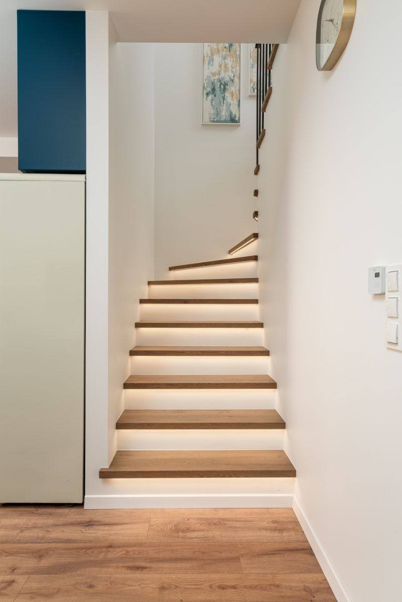 Nowoczesne schody drewniane z podświetleniem LED, białe ściany, drewniana podłoga, minimalistyczny design, zegar na ścianie, obraz w tle.