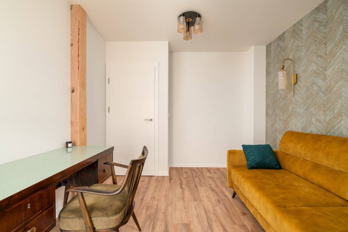 Jasne wnętrze z drewnianym biurkiem, krzesłem i musztardową sofą. Na ścianie wzorzysta tapeta i złoty kinkiet. Nowoczesna lampa sufitowa dopełnia wystrój.