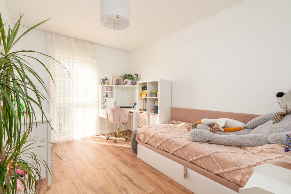 Jasny pokój dziecięcy z drewnianą podłogą, biurkiem, regałem i łóżkiem z pluszowym misiem. Delikatne zasłony w oknie wpuszczają światło. Roślina doniczkowa dodaje naturalnego akcentu.