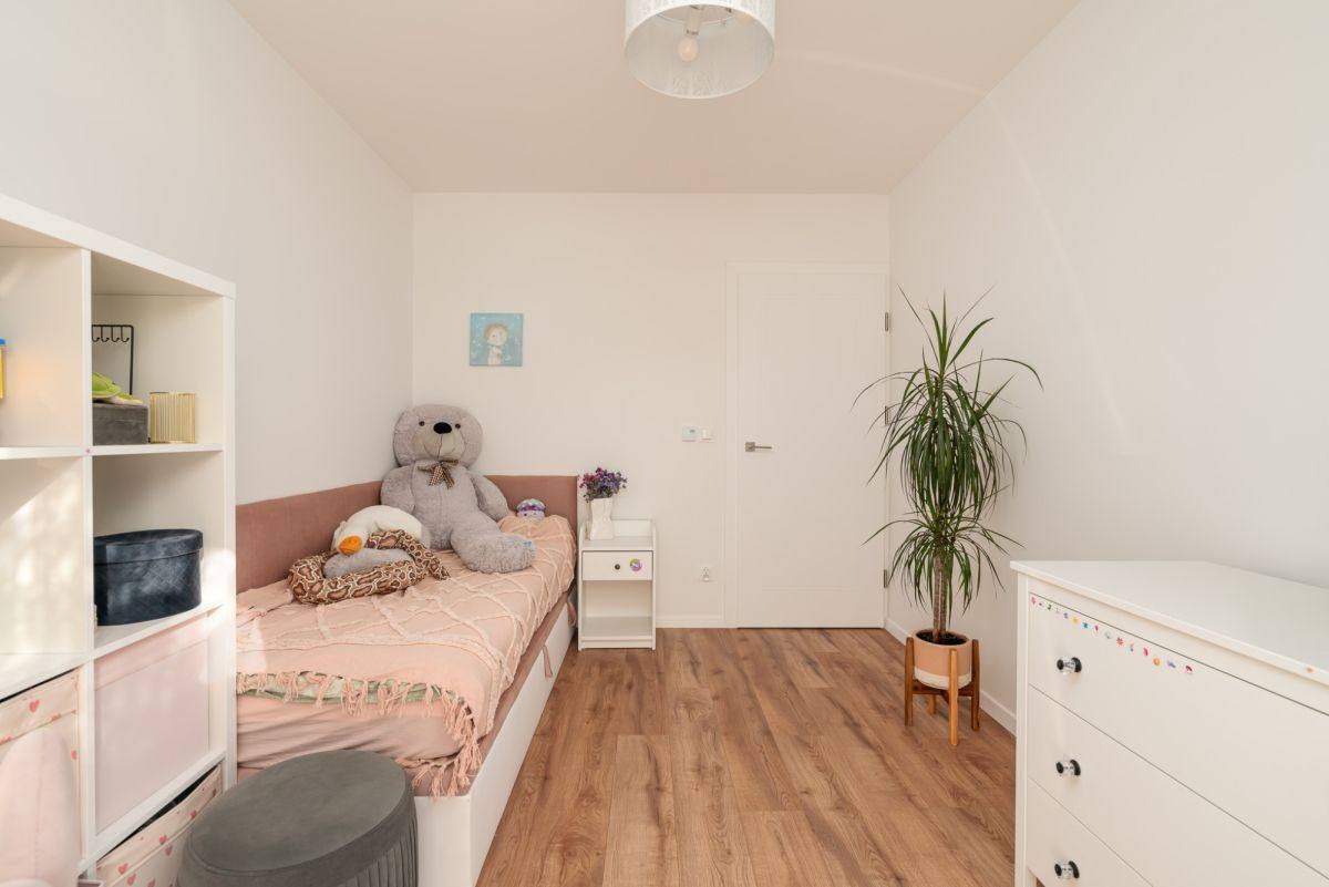 Jasny pokój dziecięcy z drewnianą podłogą. Widoczne łóżko z pluszakami, biała komoda z naklejkami, regał i roślina doniczkowa. Minimalistyczny wystrój.