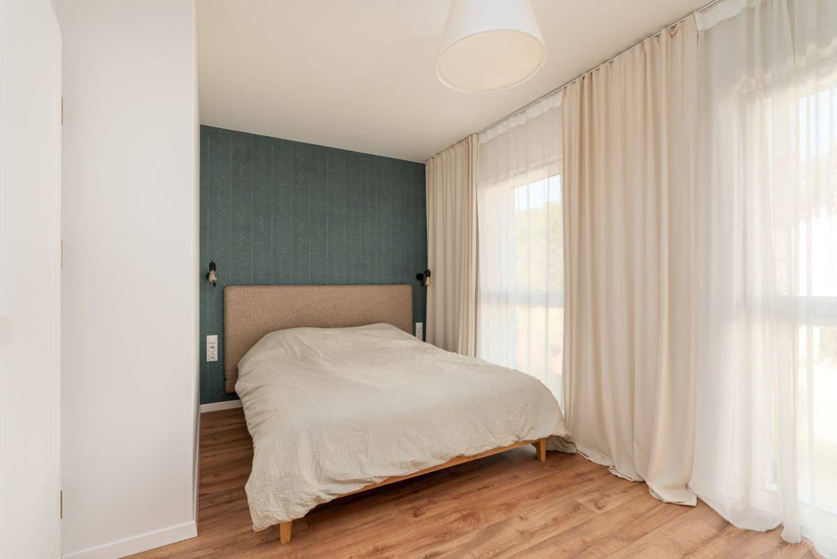 Minimalistyczna sypialnia z łóżkiem na drewnianej podłodze, zieloną ścianą i jasnymi zasłonami w oknie. Nowoczesne lampki nocne i prosta lampa sufitowa.