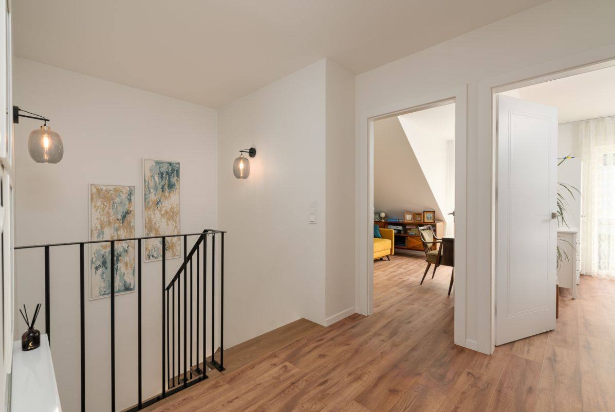 Nowoczesne wykończenie wnętrza: drewniana podłoga, czarna balustrada, abstrakcyjne obrazy i szklane kinkiety na białych ścianach, z widokiem na pokój na poddaszu.