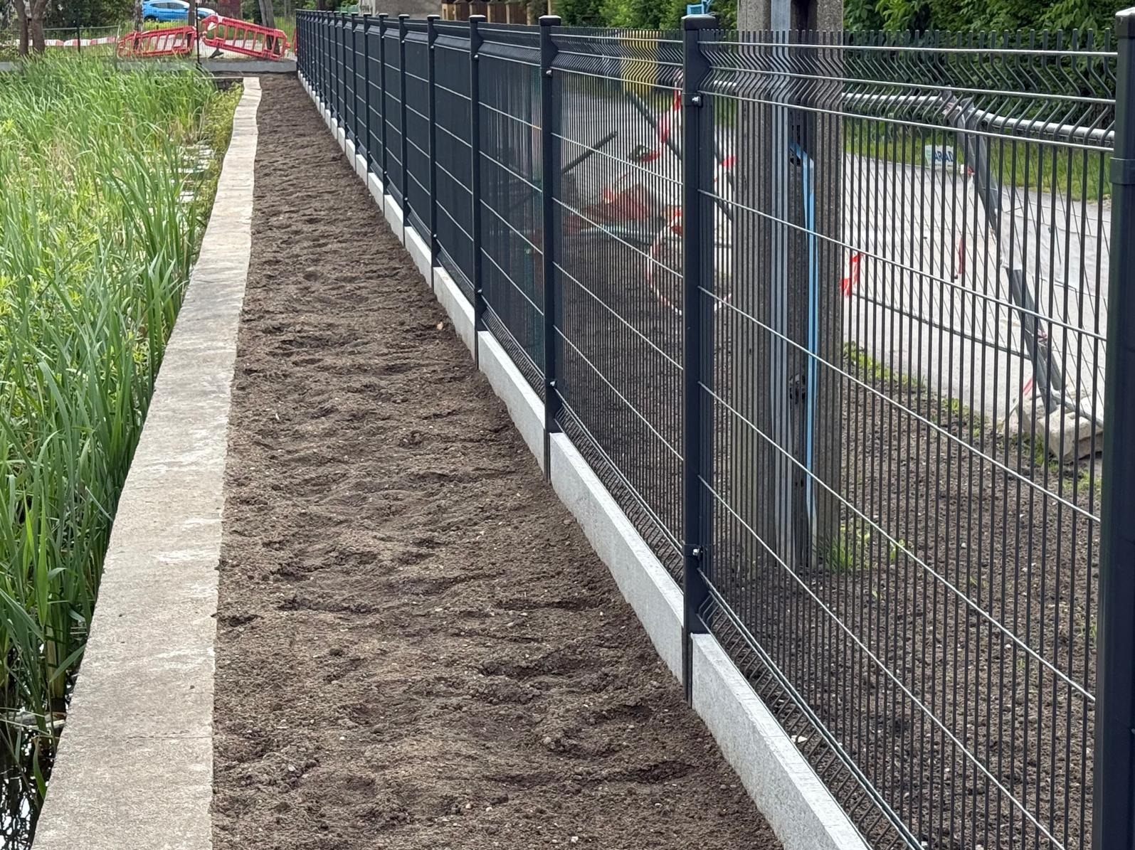 Ciemny, metalowy płot panelowy z podmurówką, widok z boku, Radwanice. Za ogrodzeniem zieleń i plac zabaw w tle. Ułożona ziemia wzdłuż ogrodzenia.