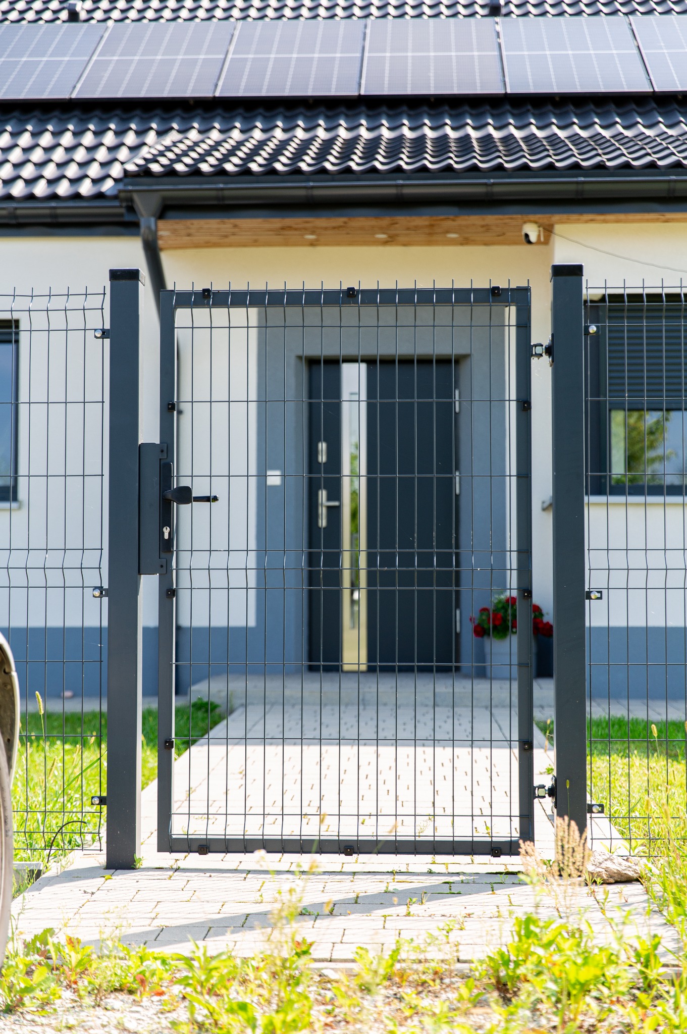 Nowoczesna, grafitowa furtka panelowa z widoczną klamką, prowadząca do domu z panelami słonecznymi na dachu. W tle donica z czerwonymi kwiatami.