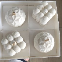 Z miłości do słodkości Magdalena Mroczkowska - Cztery białe desery musowe w pudełku, każdy z innym kształtem i złotym akcentem, trzymane przez osobę w czarno-białym swetrze na tle podłogi.