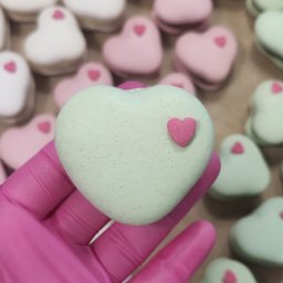 Z miłości do słodkości Magdalena Mroczkowska - Ręka w różowej rękawiczce trzyma makaronik w kształcie serca z Pyrzyc. W tle pudełko pełne makaroników w pastelowych kolorach, również w kształcie serc.