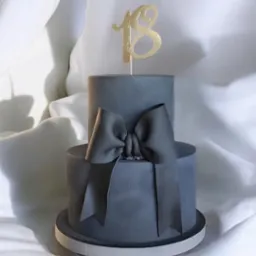 Dwupoziomowy tort w odcieniach szarości, ozdobiony kokardą i złotym topperem z liczbą 18, na tle białej, delikatnie pofałdowanej tkaniny.