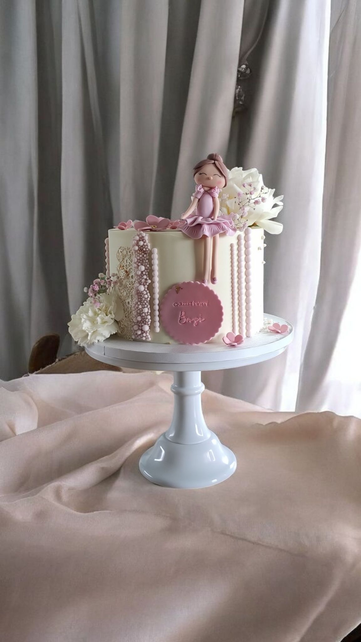 Tort na chrzest z figurką baletnicy, kwiatami i napisem 'Chrzest Święty Basi'. Kremowy tort na białej paterze, tło z delikatnych, szarych zasłon.