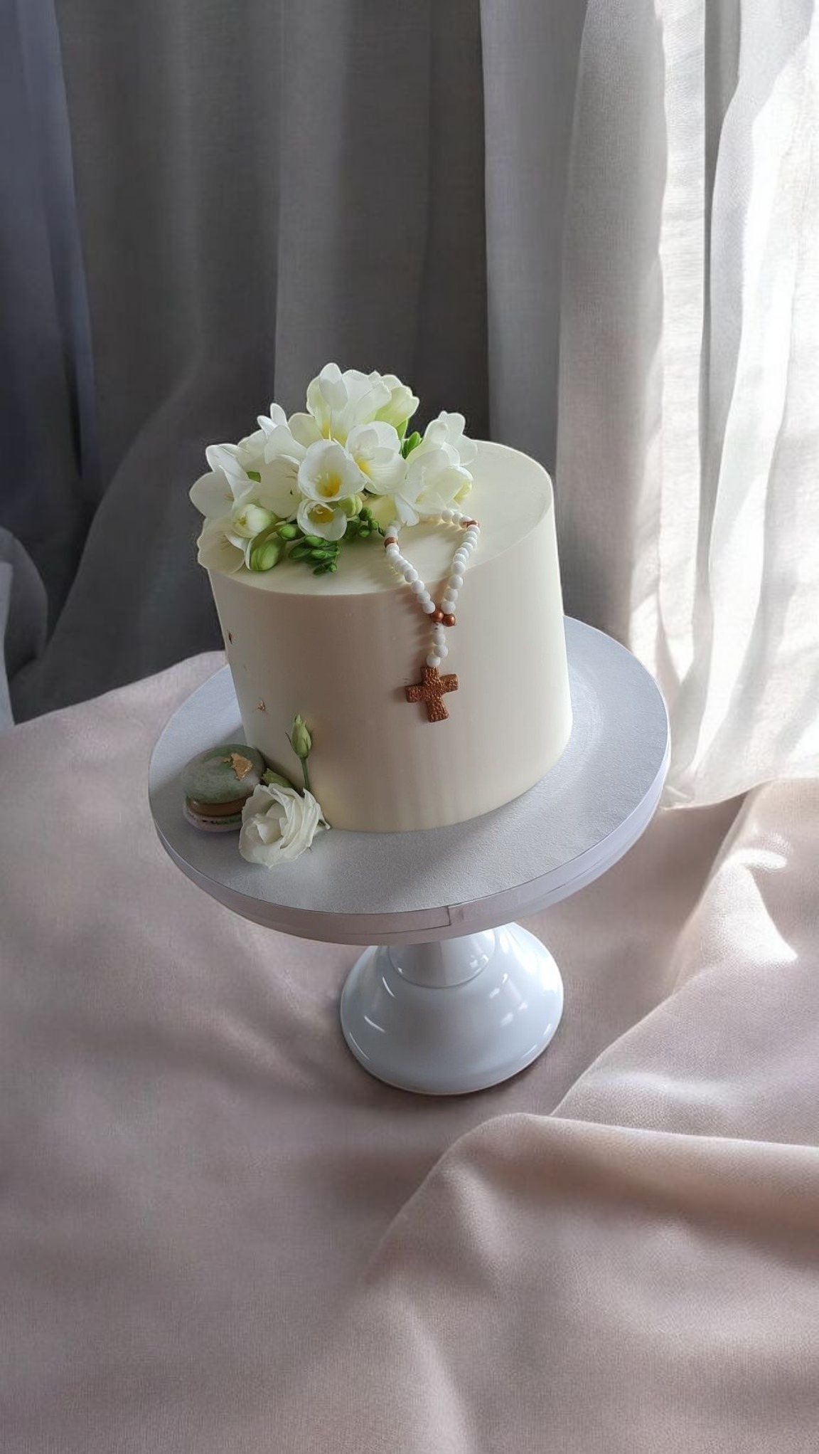 Elegancki tort komunijny na białym stojaku, zdobiony białymi kwiatami, różańcem i makaronikiem. Subtelne światło podkreśla kremową polewę.