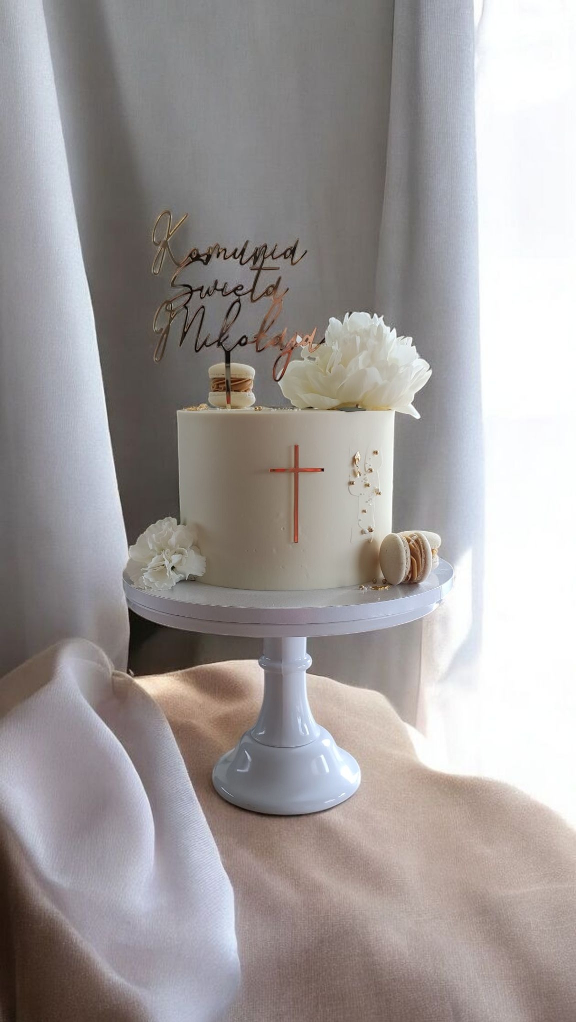 Tort komunijny na białym stojaku, zdobiony krzyżem, makaronikami i kwiatami, z topperem 'Komunia Święta Mikołaja', na tle jasnych tkanin.