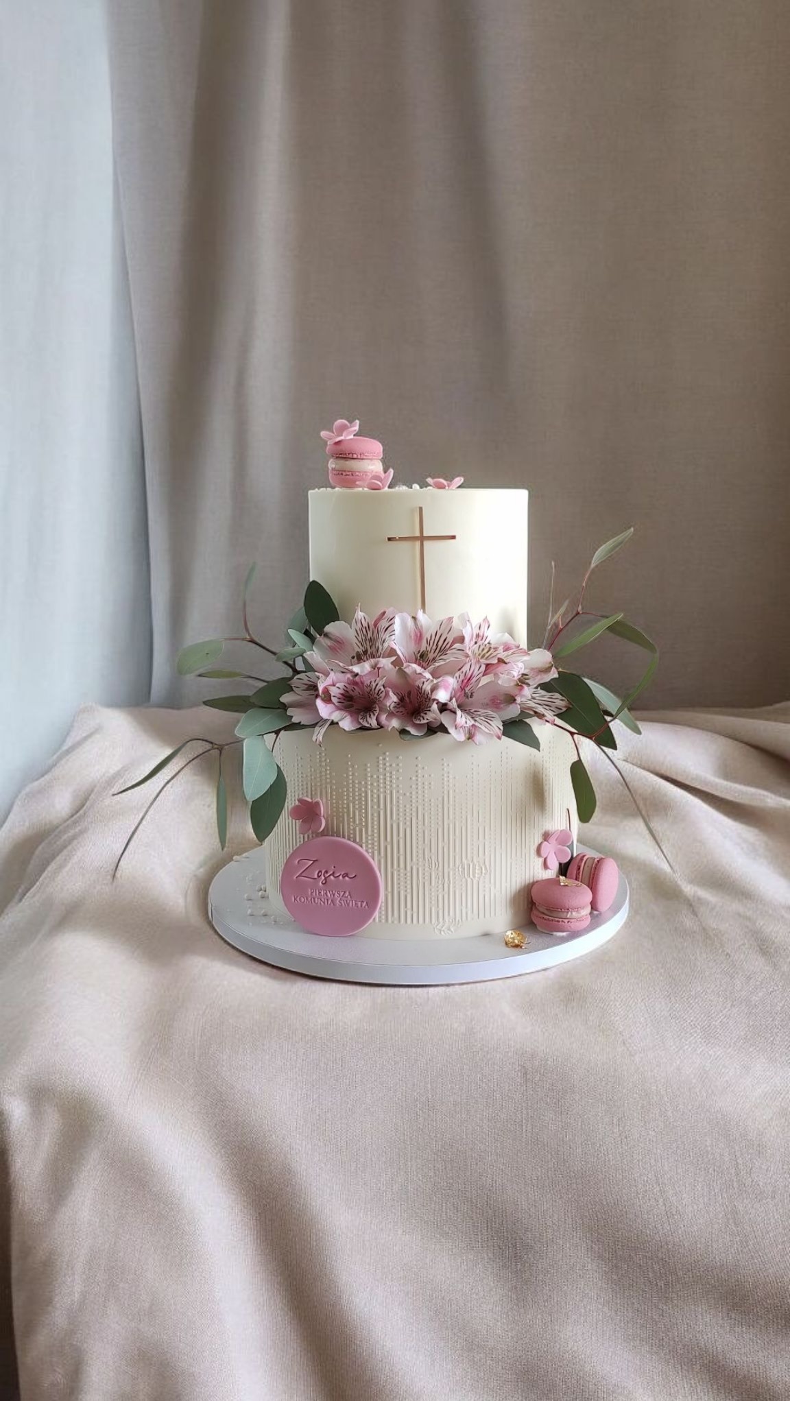 Dwupoziomowy tort komunijny w bieli, zdobiony różowymi kwiatami, makaronikami i plakietką z imieniem 'Zosia', na tle kremowej tkaniny.