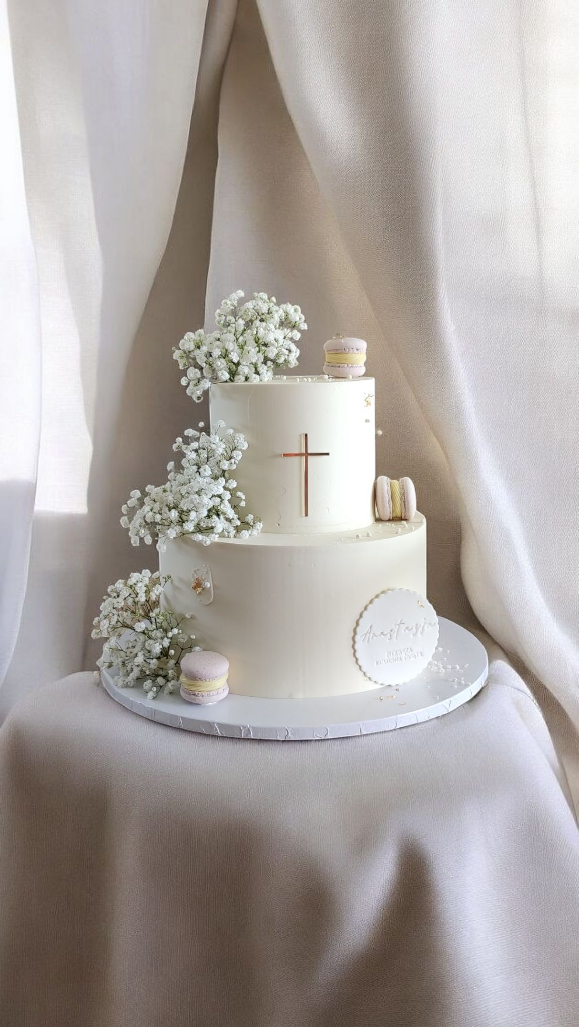 Dwupoziomowy tort w odcieniach bieli, zdobiony gipsówką, makaronikami i złotym krzyżem. Na pierwszym planie napis 'Anastazja Pierwsza Komunia Święta'.