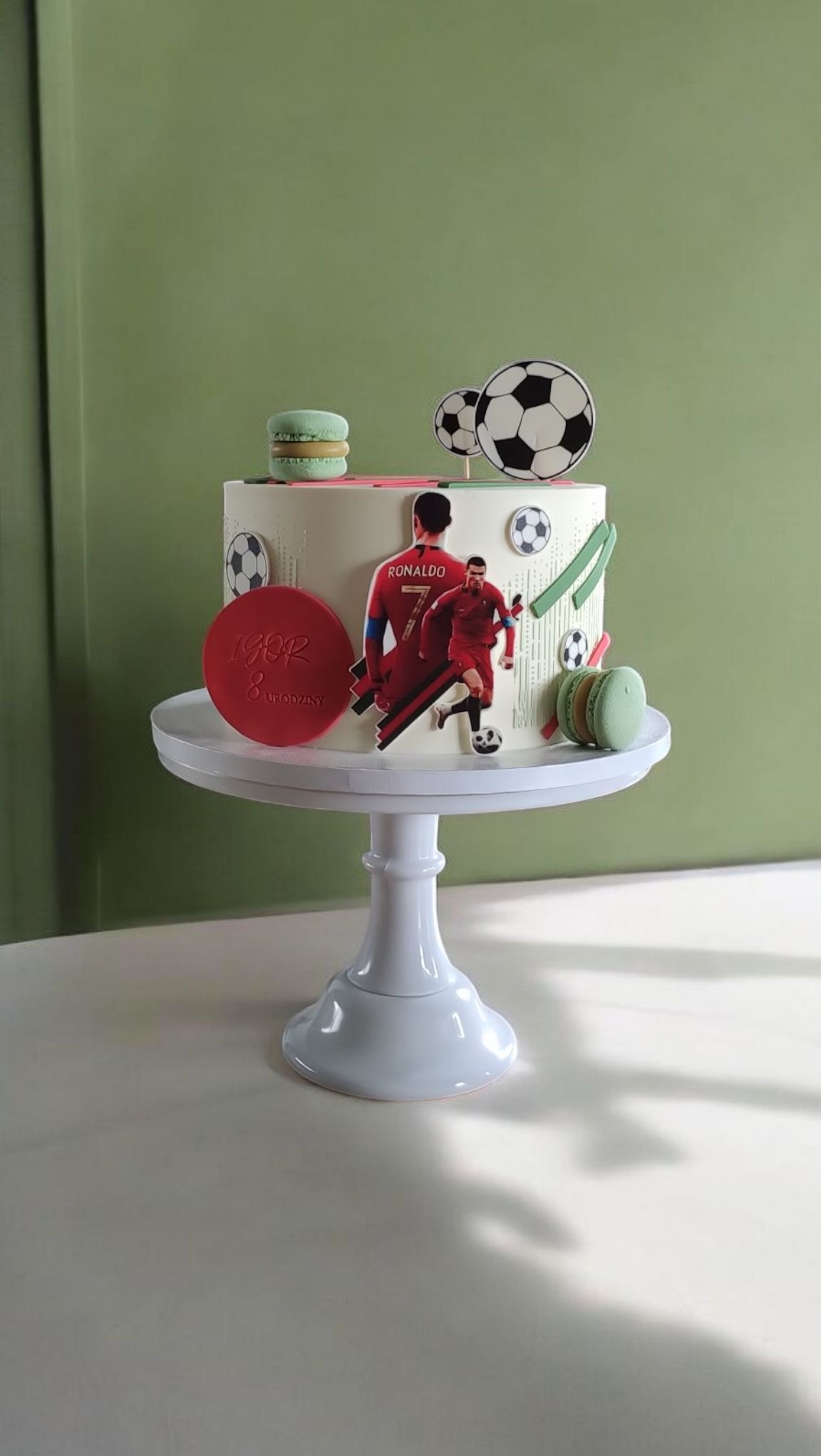 Tort urodzinowy z motywem piłkarskim, dekorowany makaronikami i wizerunkiem piłkarza na białym stojaku. Dekoracja na 8 urodziny z imieniem.