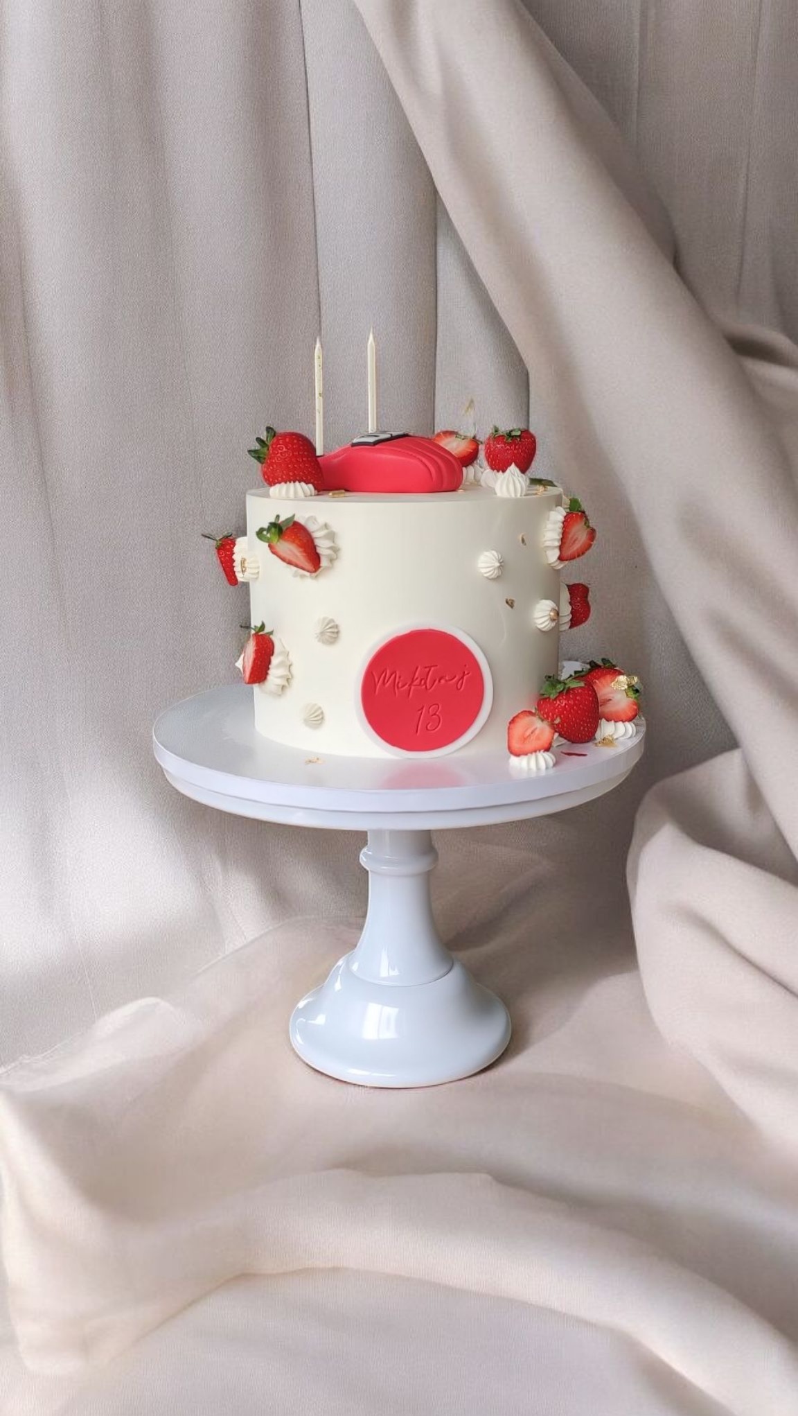 Tort urodzinowy z białym kremem, dekorowany truskawkami i czerwonym akcentem, stoi na białym, eleganckim stojaku na tle kremowej tkaniny.