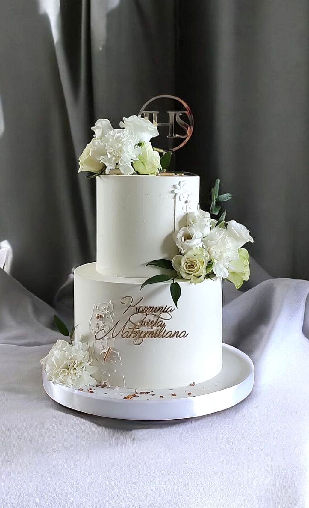Dwupoziomowy tort komunijny w bieli, zdobiony żywymi kwiatami i złotym napisem, na eleganckim tle. Subtelny, uroczysty design.