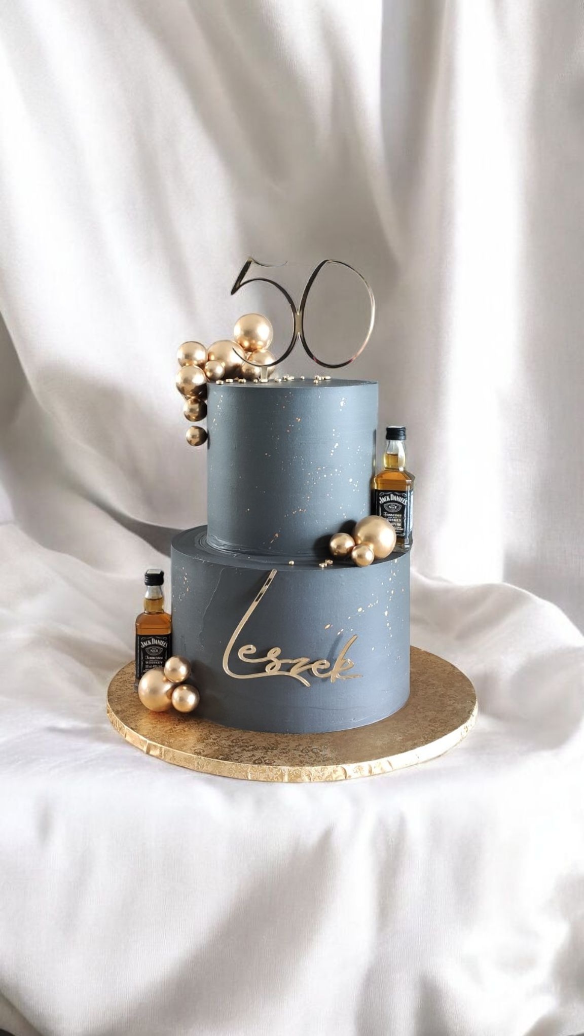 Dwupoziomowy tort w odcieniach szarości, zdobiony złotymi akcentami, mini butelkami whisky i napisem 'Leszek'. Dekoracja na 50 urodziny, na białym tle.