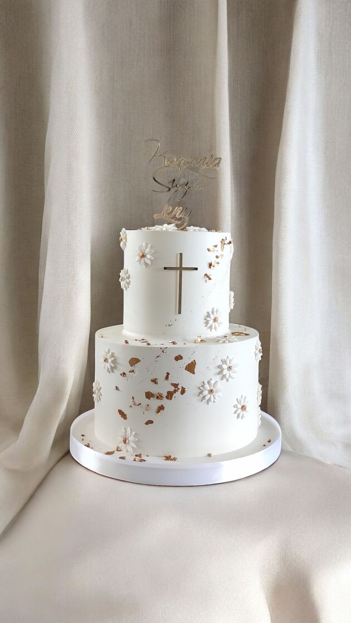 Dwupoziomowy tort komunijny w bieli, zdobiony złotymi akcentami, kwiatami i krzyżem. Na górze napis 'Komunia Święta Leny'. Tło stanowi kremowa tkanina.