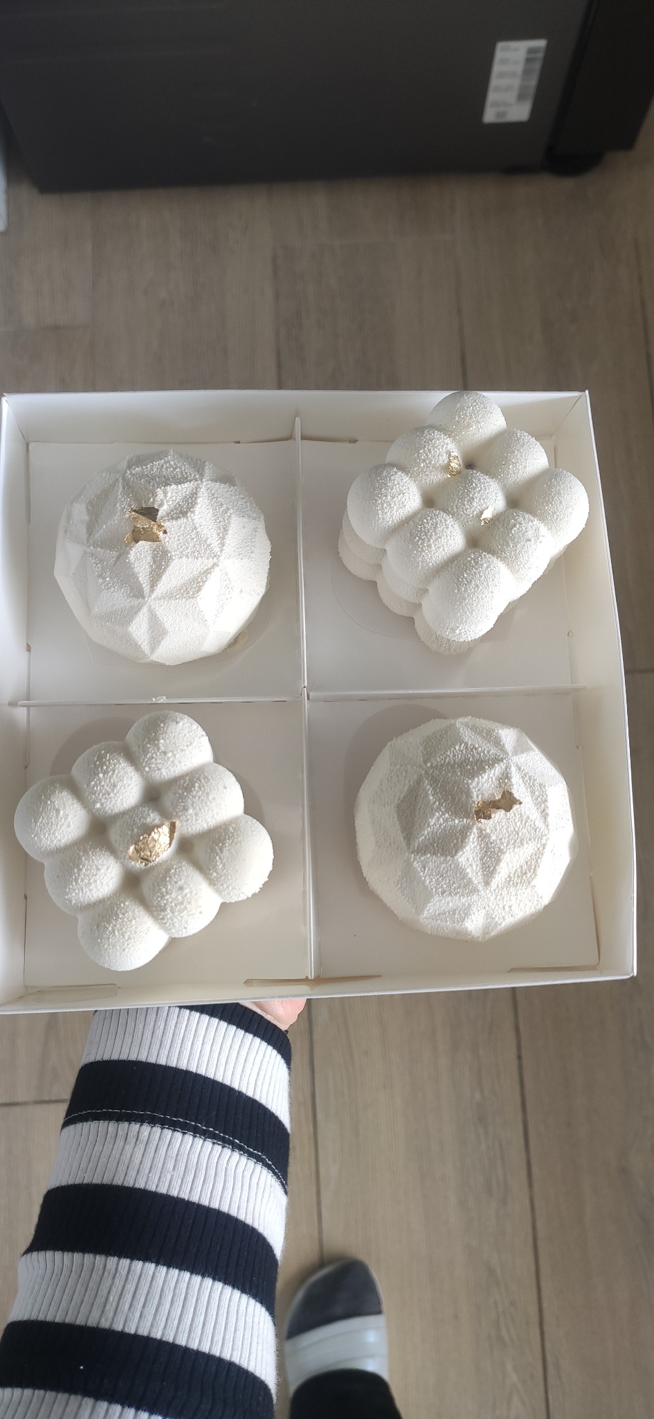 Cztery białe desery musowe w pudełku, każdy z innym kształtem i złotym akcentem, trzymane przez osobę w czarno-białym swetrze na tle podłogi.