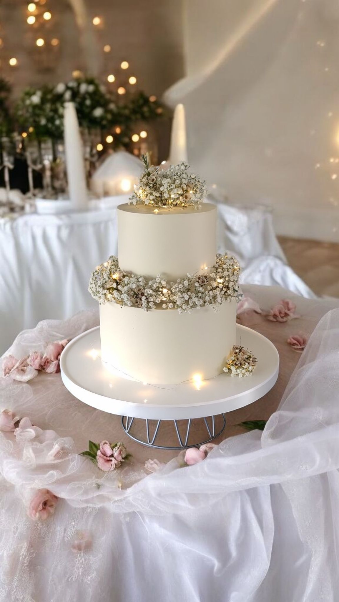 Dwupoziomowy tort w kremowym kolorze, udekorowany drobnymi kwiatami i delikatnymi światełkami, na białym stole z płatkami kwiatów i zwiewną tkaniną.