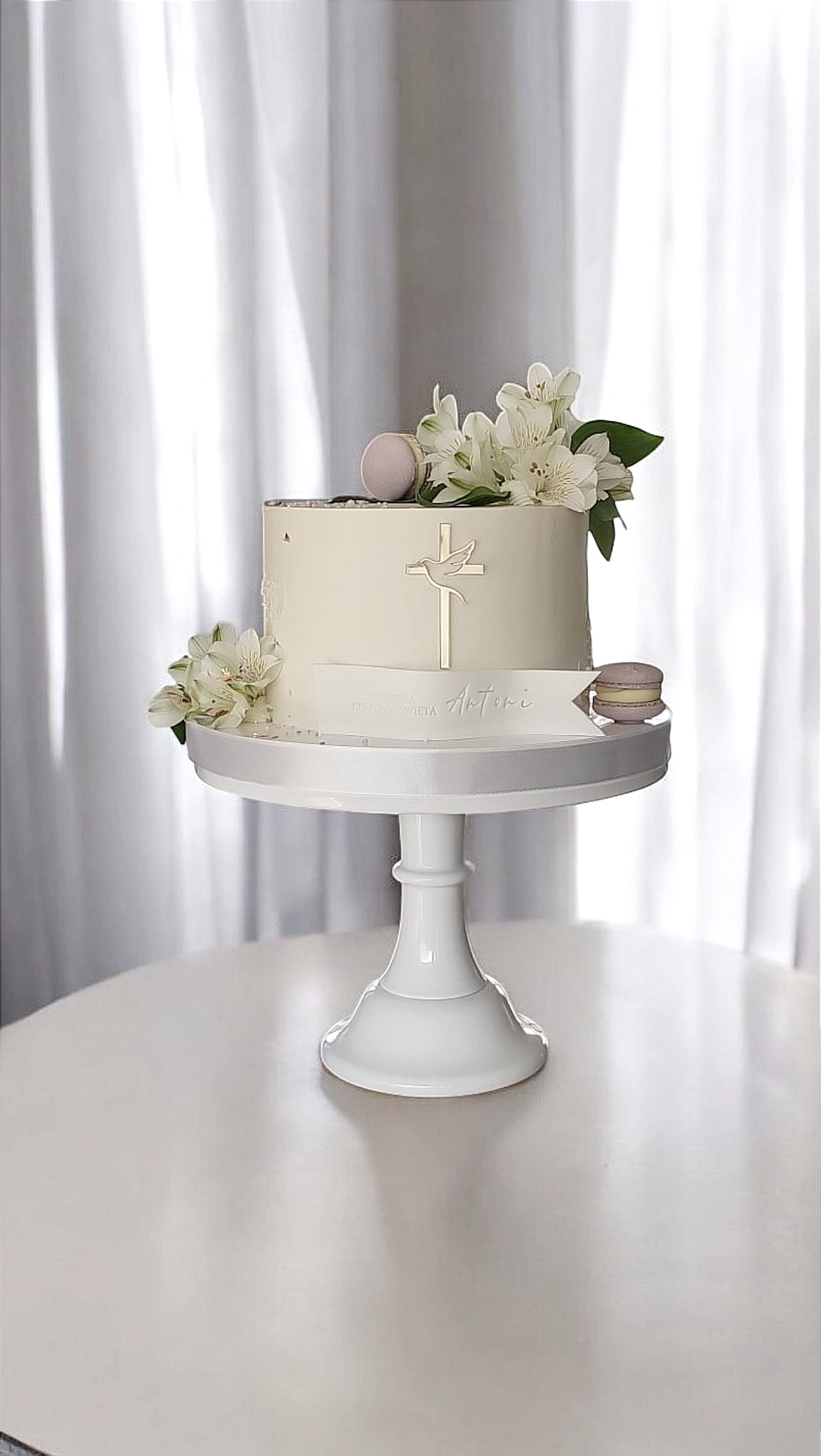 Tort komunijny w odcieniach bieli, zdobiony kwiatami, makaronikami i złotym krzyżem z gołębicą, na białej paterze, z napisem 'Pierwsza Komunia Święta Antoni'.