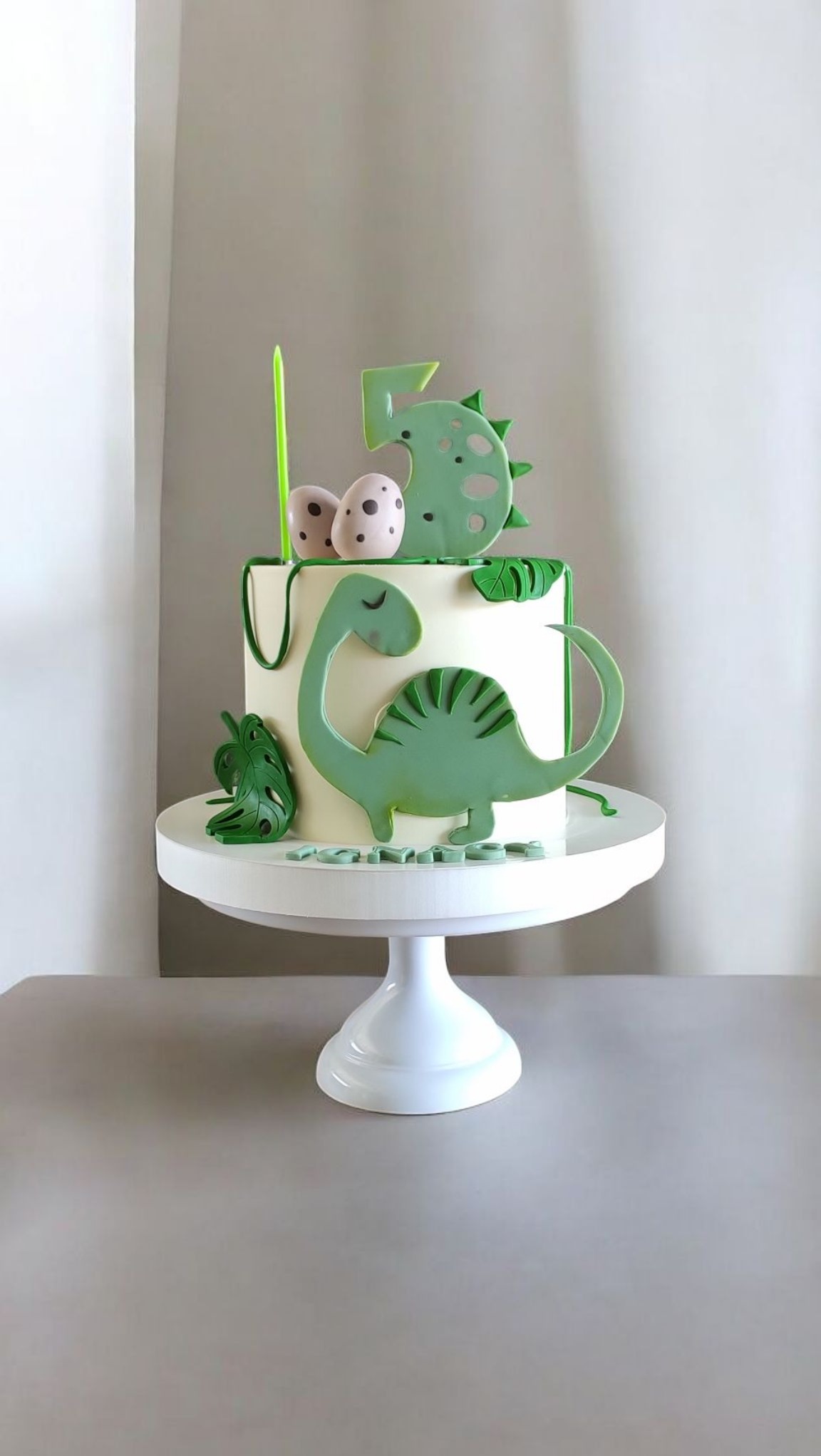 Tort urodzinowy z motywem dinozaura, dekorowany zielonymi figurkami i jajkami, umieszczony na białym stojaku na tle jasnej ściany.