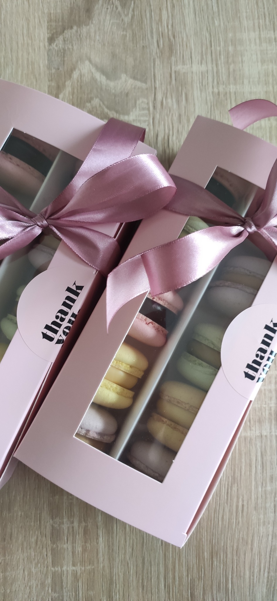Dwa pudełka z makaronikami w pastelowych kolorach, przewiązane różową wstążką. Pudełka z okienkami, na drewnianym tle, widoczne różne smaki makaroników.