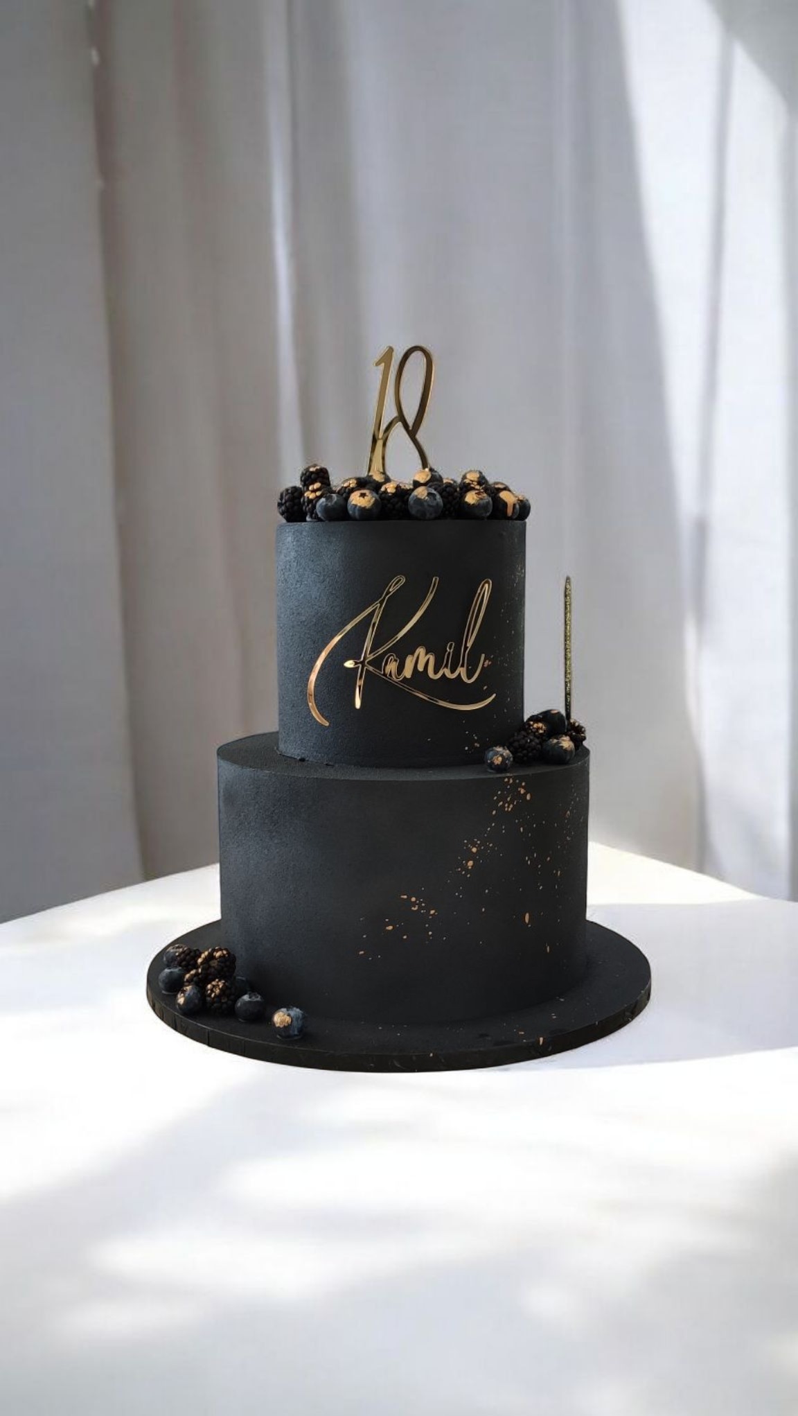 Dwupoziomowy tort w czerni, zdobiony złotymi akcentami, świeżymi jagodami i napisem 'Kamil', na białym tle. Elegancki, minimalistyczny design.