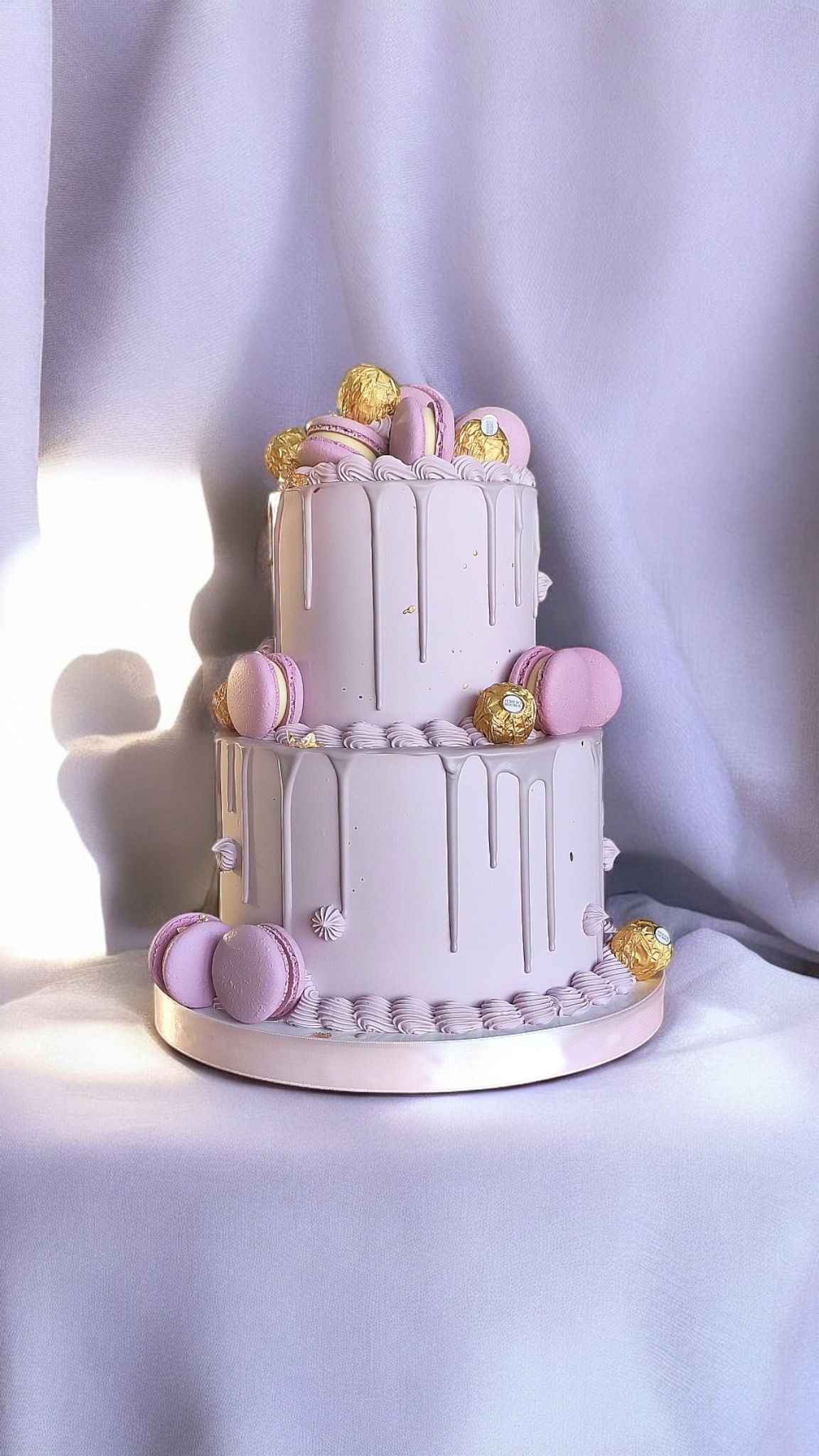 Dwupiętrowy, pudrowo-różowy tort zdobiony makaronikami i złotymi pralinkami na tle miękkiej, jasnej tkaniny. Elegancki, pastelowy deser.