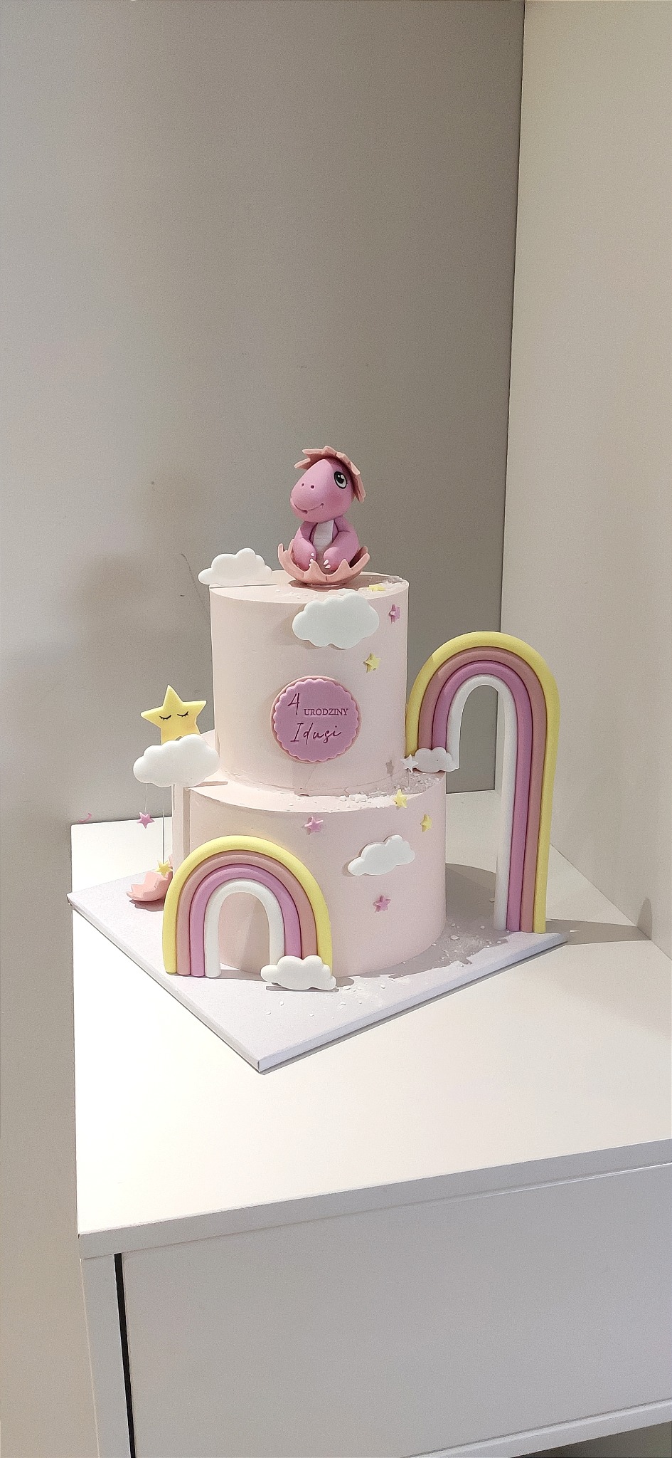 Dwupoziomowy tort urodzinowy w pastelowych kolorach z figurką różowego dinozaura, tęczami i napisem '4 Urodziny Idusi'.