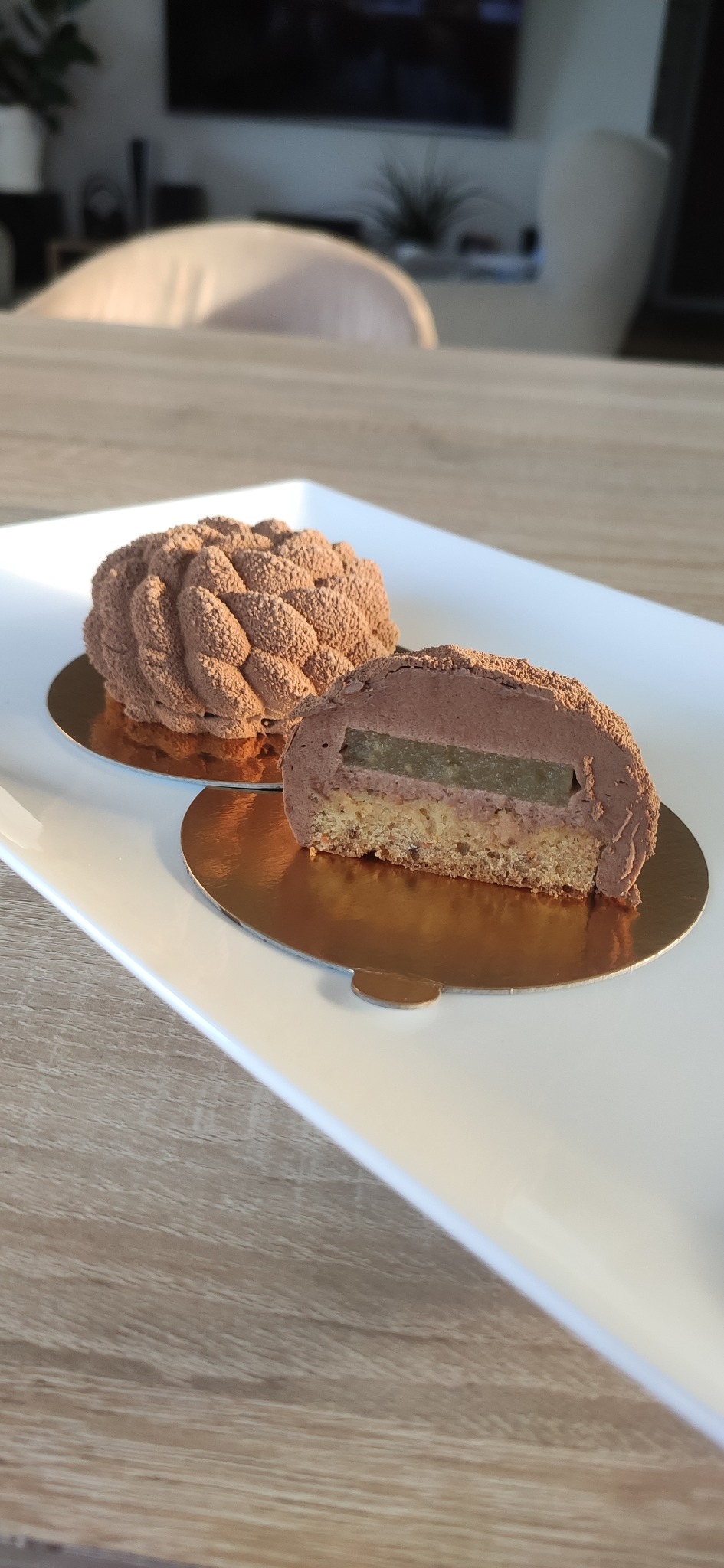 Eleganckie ciastko czekoladowe przekrojone na pół, ukazujące warstwy kremu i galaretki, obok drugiego całego ciastka na białym talerzu, na drewnianym stole.