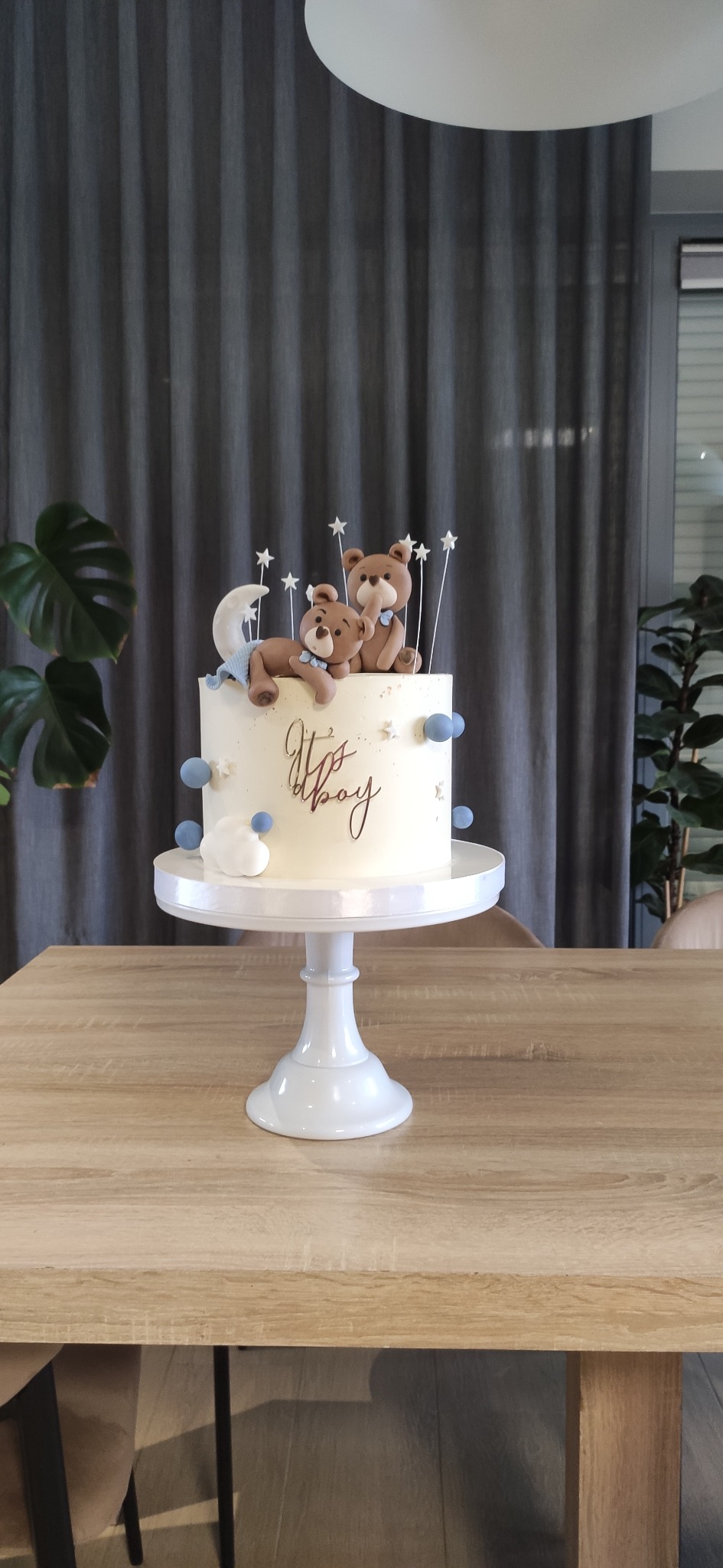 Tort 'It's a boy' na białym stojaku, zdobiony misiami, gwiazdkami i błękitnymi kulkami, na tle szarych zasłon i roślin doniczkowych. Słodki wypiek na specjalną okazję.
