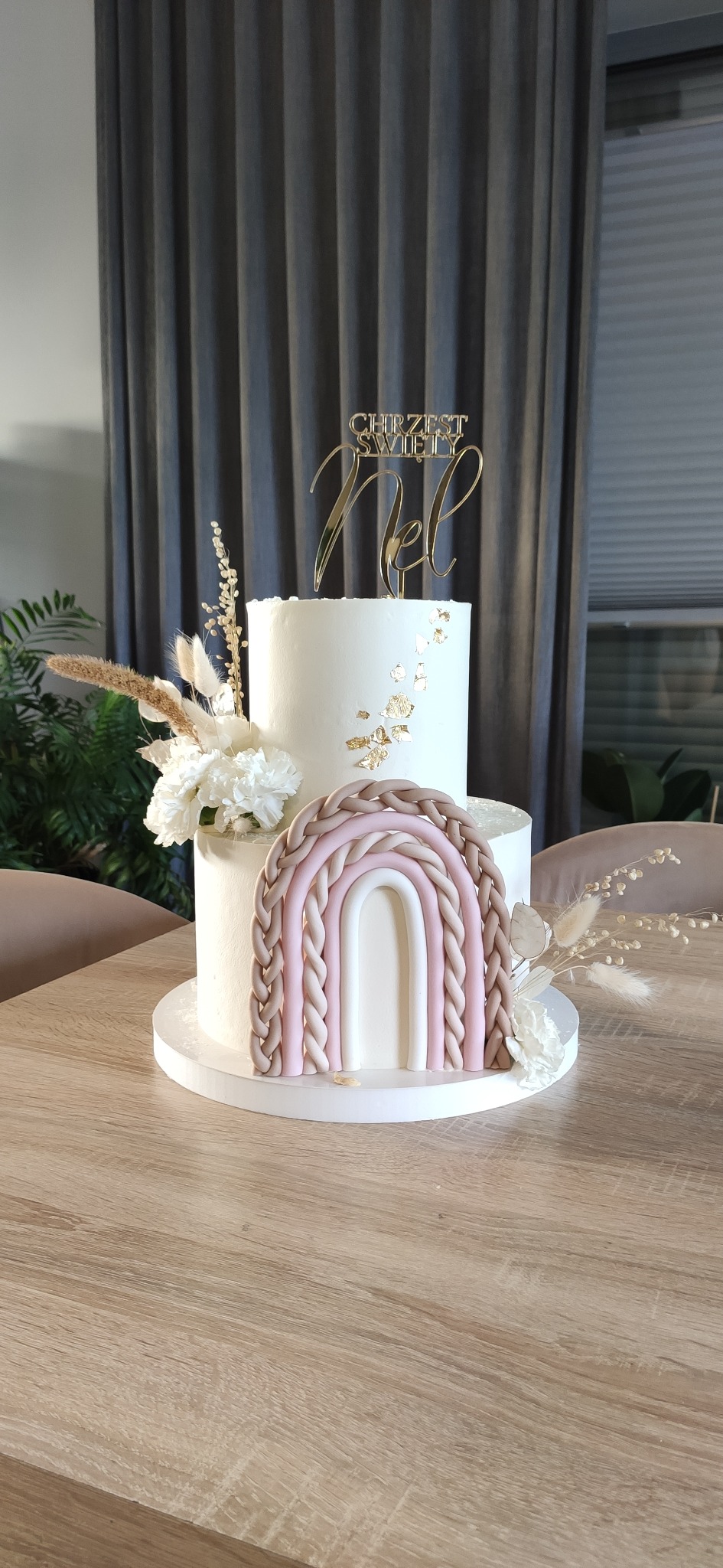 Dwupoziomowy tort na Chrzest Święty z dekoracją w postaci plecionego tęczowego łuku, złotych płatków i napisu. W tle zasłona i roślina.
