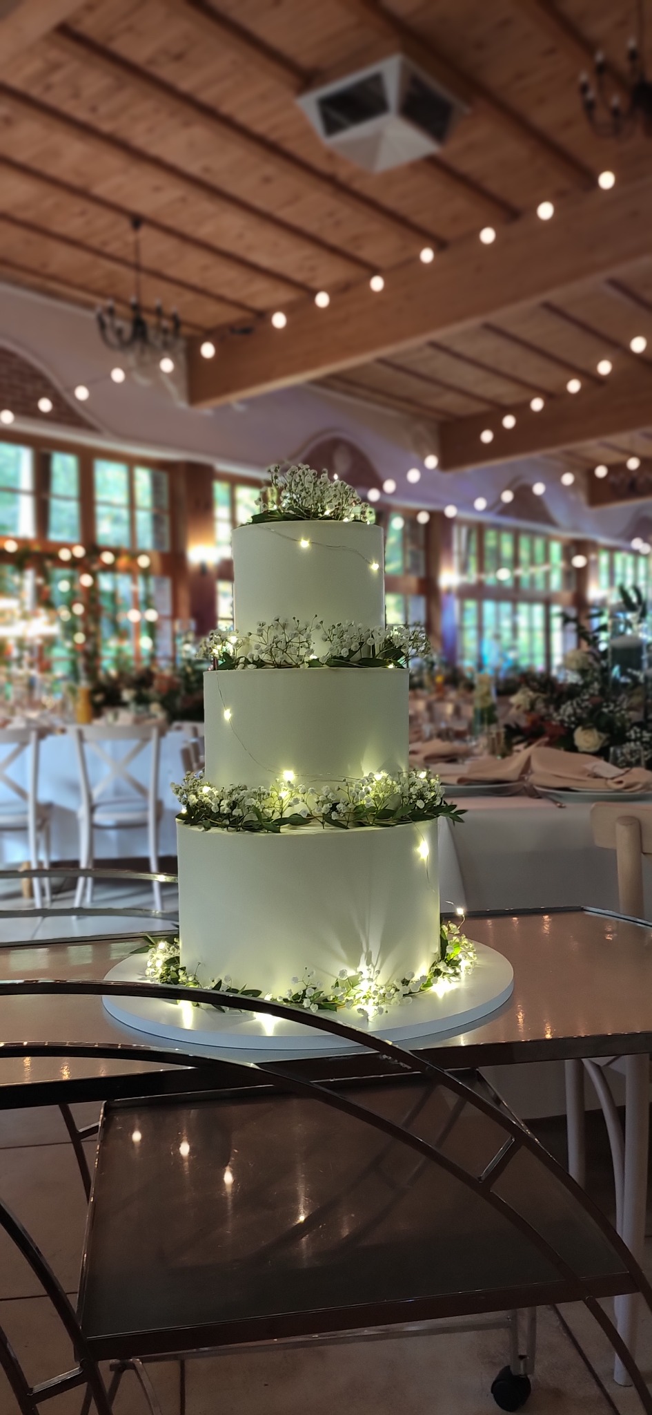 Trzypiętrowy tort weselny z białego kremu, zdobiony drobnymi kwiatami i światełkami, na tle sali bankietowej z dekoracjami świetlnymi.