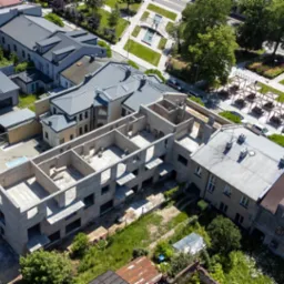 Zdjęcie z lotu ptaka przedstawia budynek w trakcie murowania ścian, z widocznymi elementami konstrukcyjnymi i otaczającą zielenią.