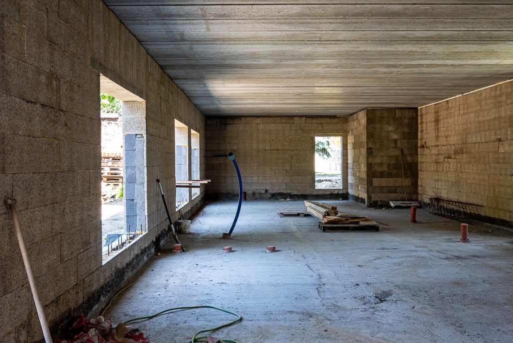 Wnętrze domu w trakcie budowy: surowe ściany z betonu komórkowego z otworami okiennymi, betonowy strop i podłoga. Widoczne narzędzia i materiały budowlane.