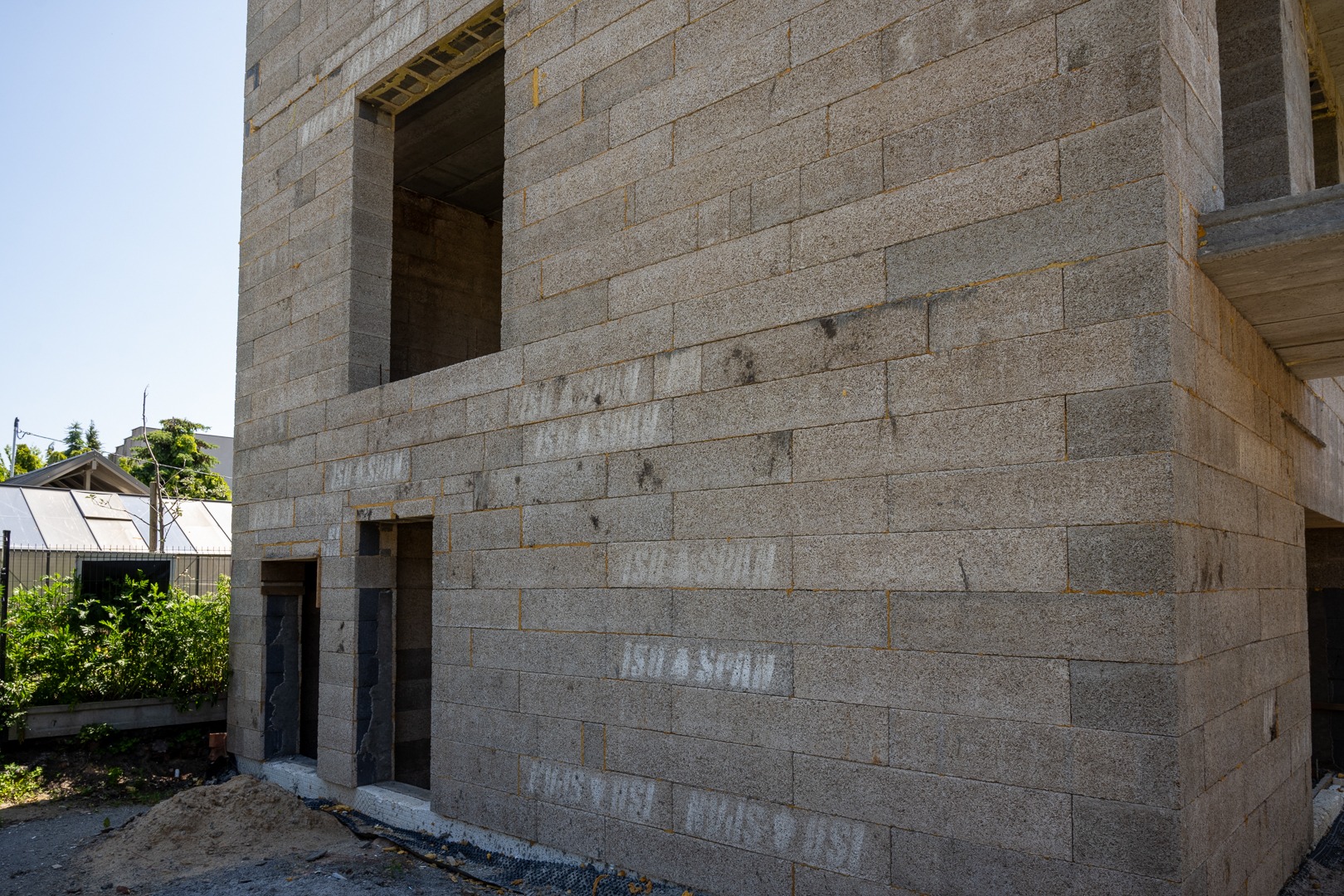 Surowa bryła domu w trakcie budowy, ściany z szarego bloczka, widoczne otwory okienne i drzwiowe. Częściowo zarośnięty teren wokół budynku.