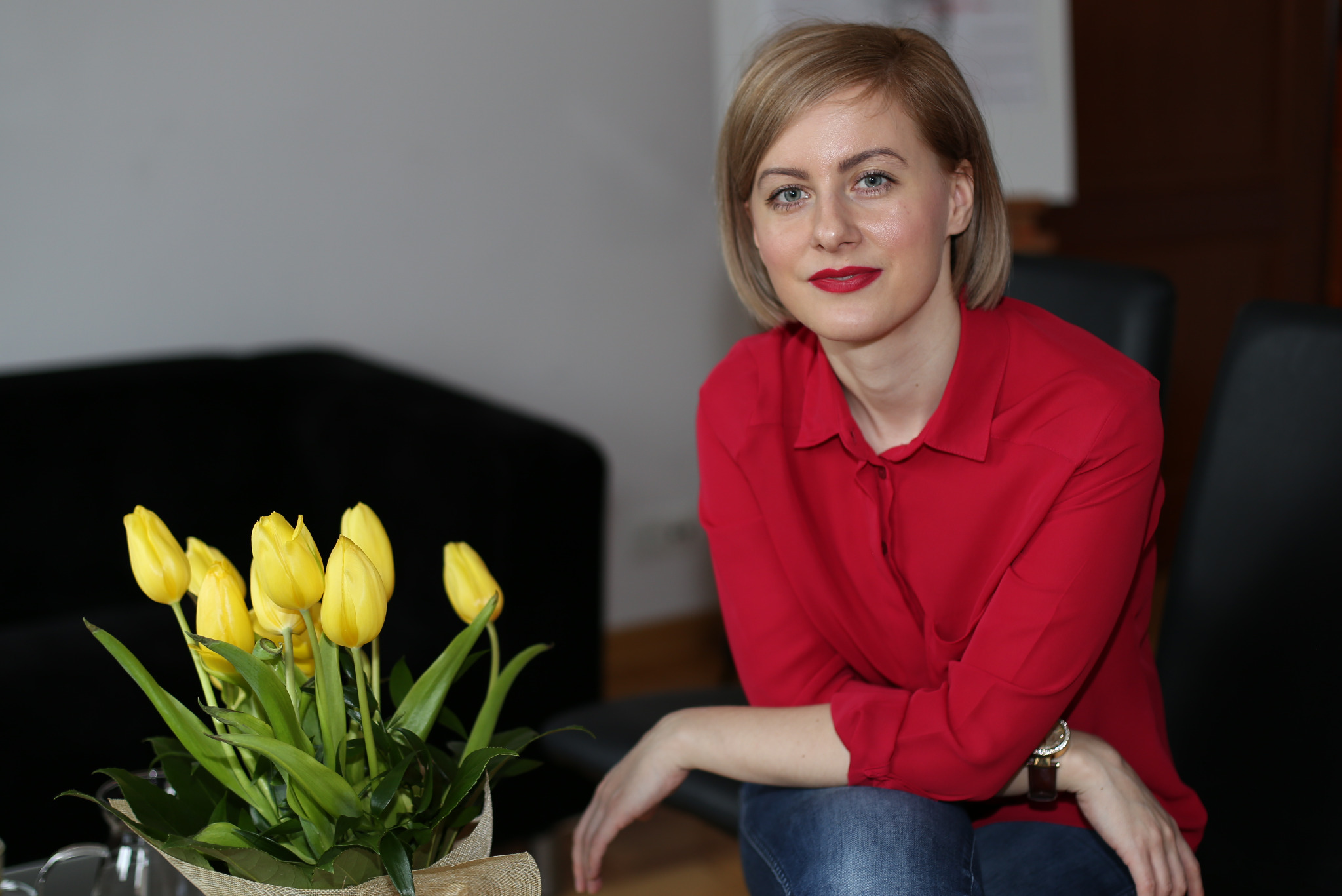 Kobieta w czerwonej koszuli siedzi obok bukietu żółtych tulipanów. Portret w naturalnym świetle, z delikatnym uśmiechem i wyraźnym makijażem.