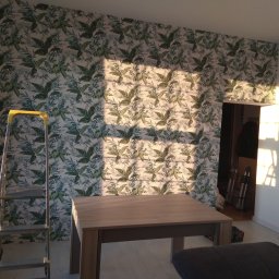 Aleksander Mielnicki - Pokój po tapetowaniu w roślinny wzór. Żółta drabina stoi przy ścianie, a na pierwszym planie stół i fragment sofy. Na ścianie widoczne promienie słoneczne.