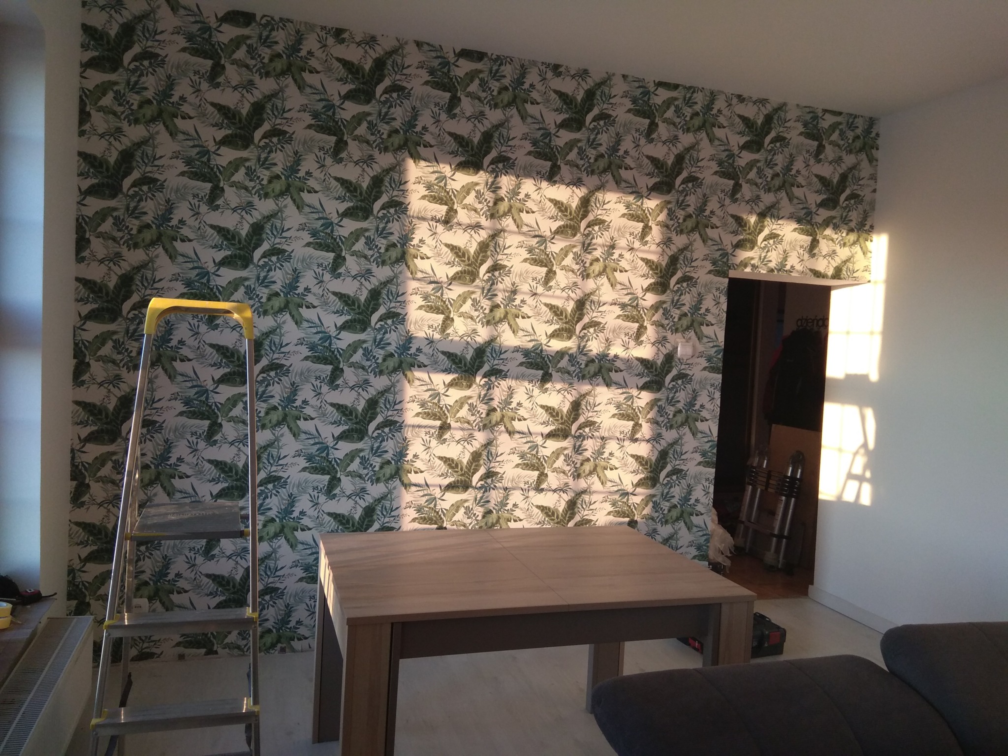 Pokój po tapetowaniu w roślinny wzór. Żółta drabina stoi przy ścianie, a na pierwszym planie stół i fragment sofy. Na ścianie widoczne promienie słoneczne.