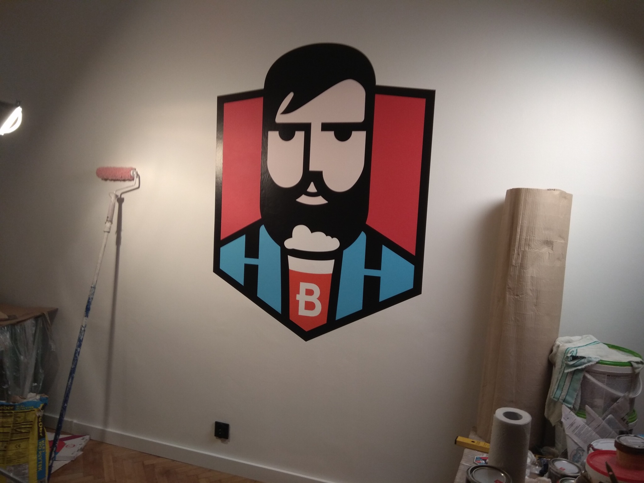 Ściana z namalowanym, kolorowym logo brodatego mężczyzny i narzędziami malarskimi. Widoczny wałek do malowania i karton zabezpieczający podłoże.