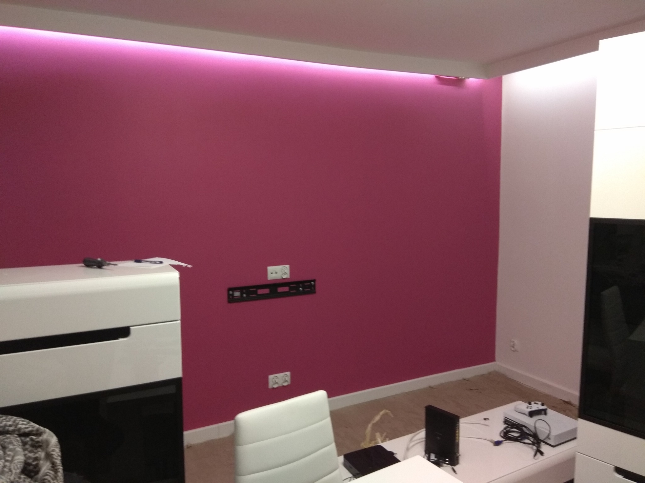 Pokój po remoncie: ściana w kolorze fuksji z podświetleniem LED, zamontowany uchwyt TV, białe meble. Widoczne gniazdka elektryczne i sprzęt RTV.