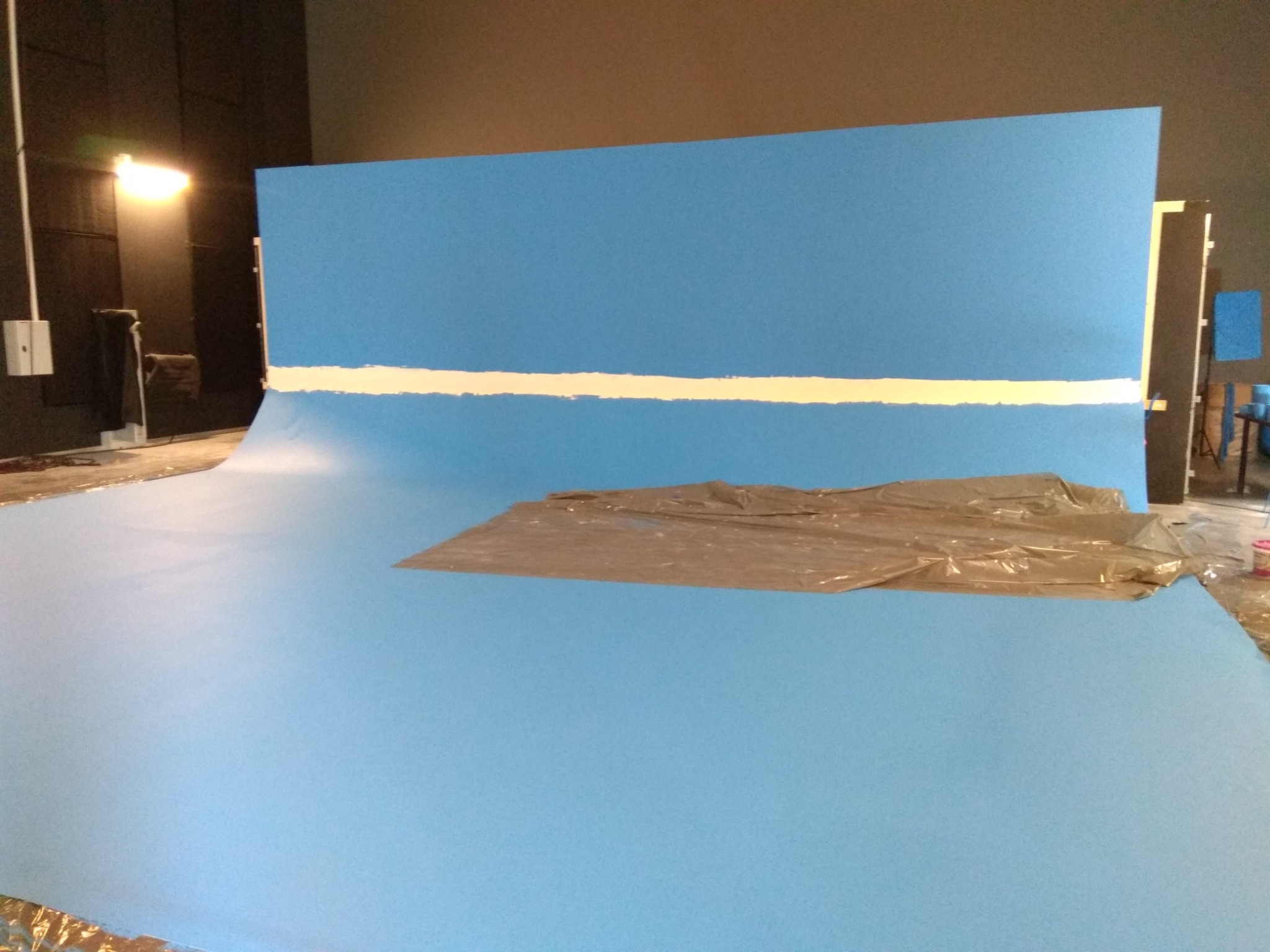 Przygotowanie studia malarskiego: niebieskie tło i podłoga, zabezpieczona folią. Biała linia na ścianie, lampa oświetleniowa w tle. Etap prac przed malowaniem.