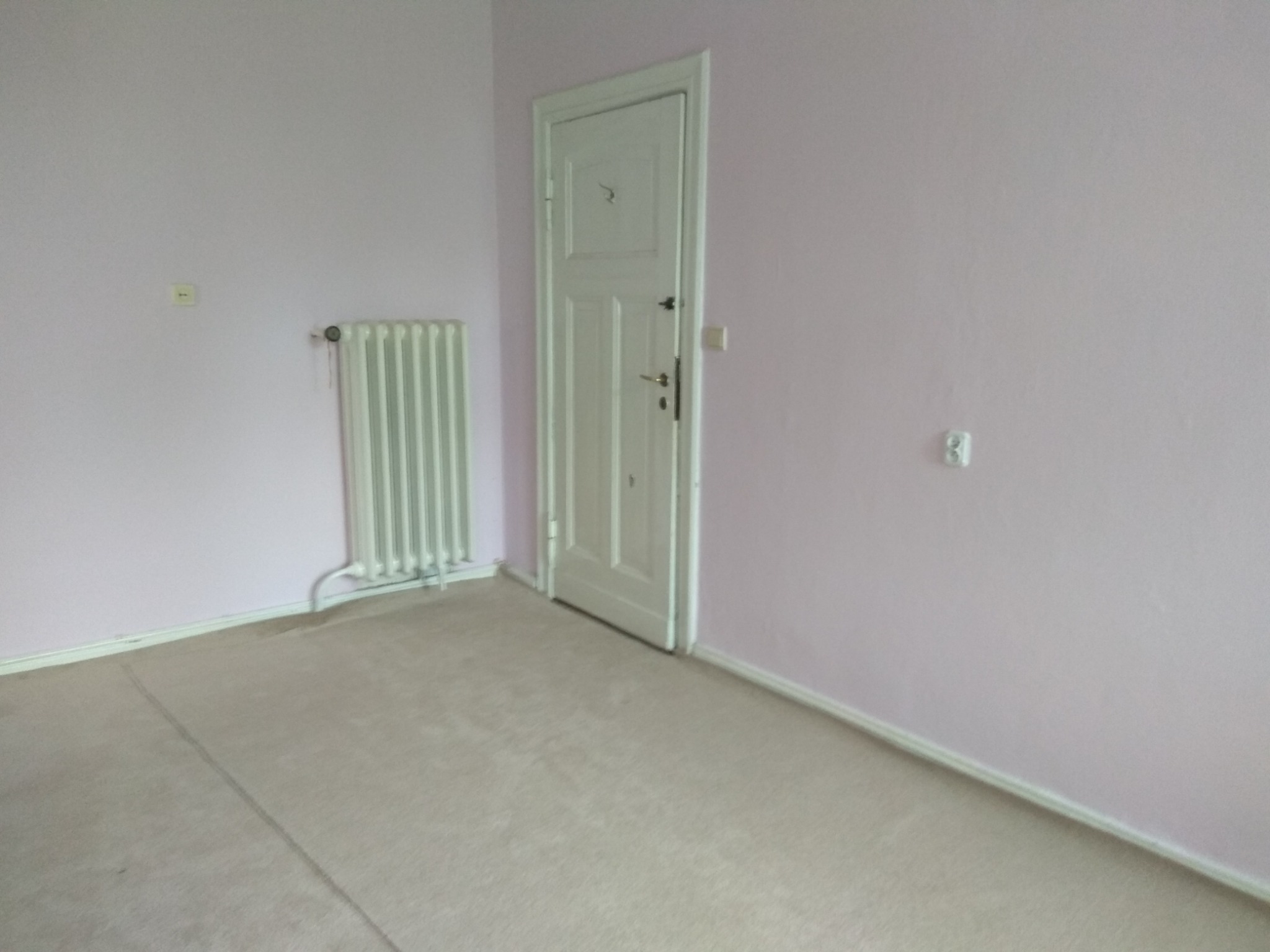 Pusty pokój w odcieniach różu z białymi drzwiami i grzejnikiem. Jasna podłoga z paneli. Widoczne gniazdko elektryczne i termostat na ścianie. Delikatne światło dzienne.