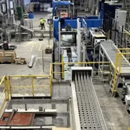 Wnętrze hali produkcyjnej z suwnicą, linia produkcyjna profili stalowych, pracownik w odblaskowej kamizelce. Wysoka perspektywa, widok na proces obróbki metalu.