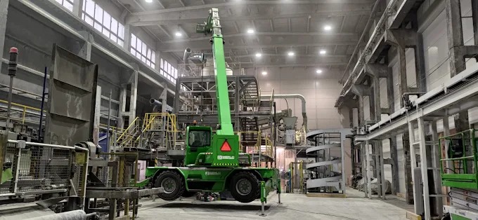 Wnętrze hali produkcyjnej z suwnicą teleskopową Merlo w kolorze zielonym, podnoszącą element konstrukcyjny. Widoczne elementy konstrukcyjne hali i oświetlenie.