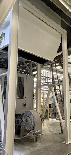 Wnętrze hali z konstrukcją stalową, widoczne elementy maszyny i drabina. Ujęcie z dołu, neutralne oświetlenie.